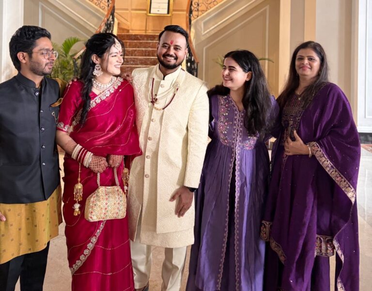 This image captures a joyful group of five people standing together, likely at a wedding or festive celebration. At the center, a bride and groom are dressed in traditional Indian attire. The bride is wearing a rich red saree adorned with gold embroidery, heavy jewelry, and bangles, and is holding an ornate handbag. The groom is dressed in a cream-colored sherwani with subtle patterns and a red tilak on his forehead. They are flanked by three guests: a man in a black and mustard kurta on the left, and two women in matching purple ethnic outfits with intricate embroidery on the right. All of them are smiling warmly, looking at each other and clearly enjoying the occasion. The background features a staircase and elegant interior décor, enhancing the festive and grand setting.