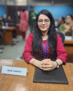 This image shows a woman seated at a wooden desk in a formal setting, likely a conference or training session. She is wearing a maroon traditional outfit with subtle embroidery and has long black hair. She is also wearing glasses and a blue lanyard around her neck, suggesting she is a participant or delegate. Her hands are neatly folded on a black folder placed on the desk in front of her. A nameplate on the table clearly displays the name “Jyoti Lalwani.” The background shows other people seated and interacting, with the focus softly blurred to emphasize Jyoti in the foreground. She is smiling calmly, creating a composed and confident presence.