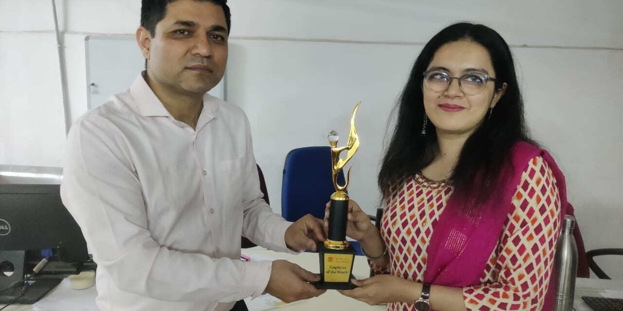 This image shows a man and a woman standing together in what appears to be an office environment. The man, dressed in a light-colored formal shirt and dark trousers, is handing a trophy to the woman. The woman, wearing glasses and a traditional outfit with a cream and red patterned design paired with a magenta dupatta (scarf), is smiling as she accepts the award. The trophy is gold and black with a flame-like design on top, and the label on it reads "Employee of the Month." They are standing beside a desk with office items like a computer, files, and a water bottle. The atmosphere suggests a recognition or award ceremony.