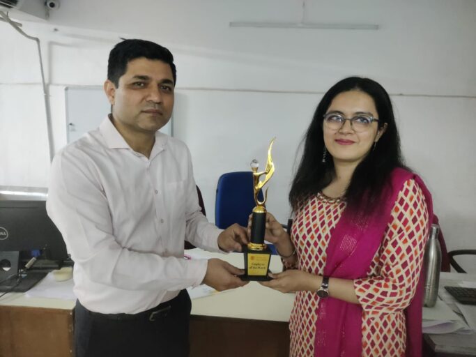This image shows a man and a woman standing together in what appears to be an office environment. The man, dressed in a light-colored formal shirt and dark trousers, is handing a trophy to the woman. The woman, wearing glasses and a traditional outfit with a cream and red patterned design paired with a magenta dupatta (scarf), is smiling as she accepts the award. The trophy is gold and black with a flame-like design on top, and the label on it reads "Employee of the Month." They are standing beside a desk with office items like a computer, files, and a water bottle. The atmosphere suggests a recognition or award ceremony.